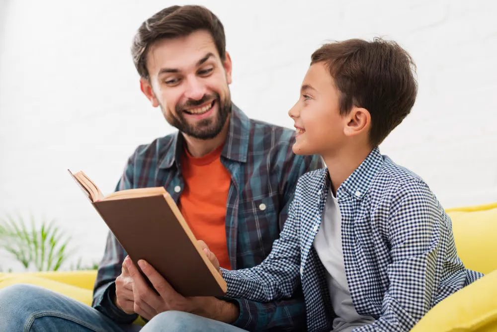 A father and son sitting on a sofa reading a book together.