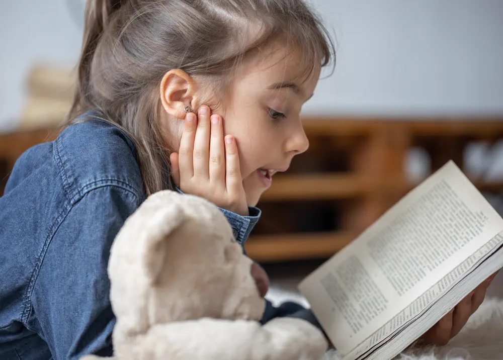 Young girl reading a book, chin in hand, beside a teddy bear